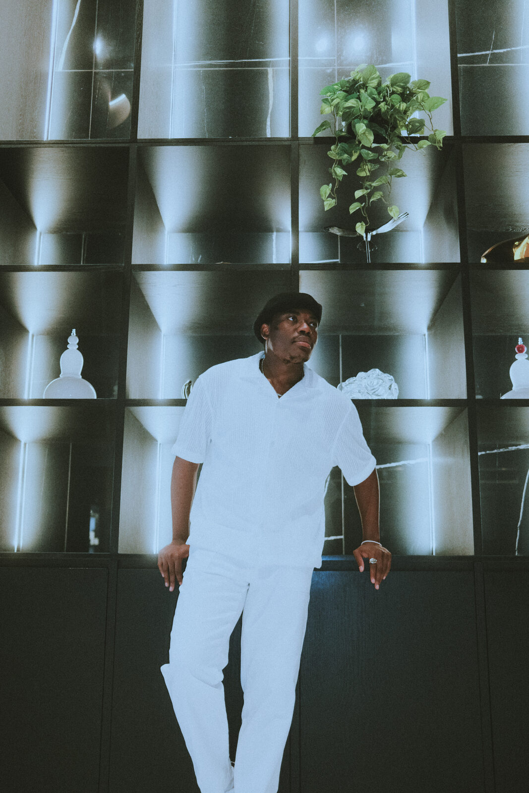 Elvic Kongolo dressed in white, standing in front of a lit shelf wall