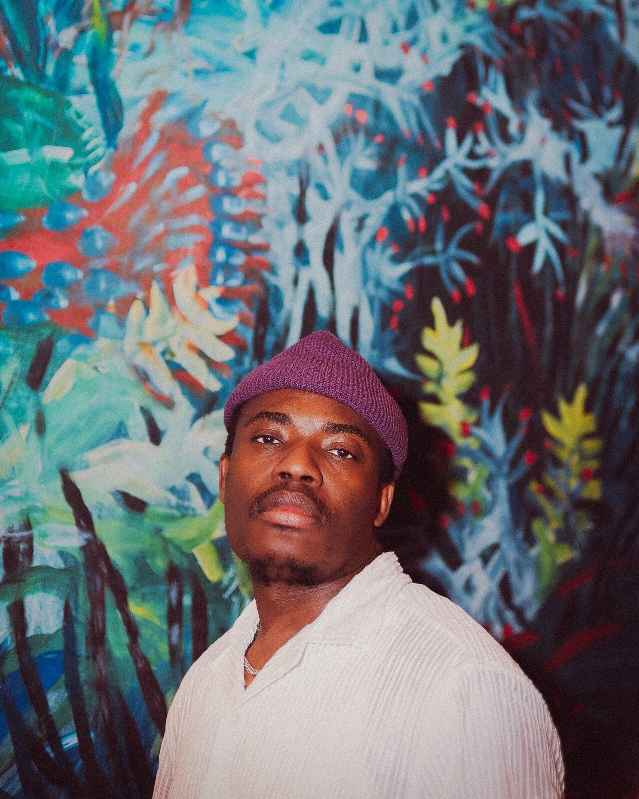 Portrait of Elvic Kongolo in a purple beanie and white shirt in front of a colourful painted jungle mural